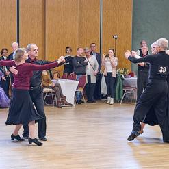 Zwei Paare tanzen in eleganter Kleidung auf einer Tanzfl&auml;che, w&auml;hrend eine Zuschauergruppe beobachtet.