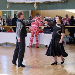Tanzpaar f&uuml;hrt einen langsamen Walzer auf einer Tanzfl&auml;che, Zuschauer im Hintergrund, festliche Stimmung im Saal.