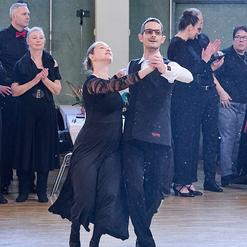 Tanzpaar in eleganter Kleidung f&uuml;hrt einen Schritt auf der Tanzfl&auml;che, umgeben von applaudierenden Zuschauern.