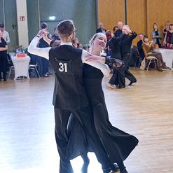 Tanzpaar in festlicher Kleidung tanzt auf einer Tanzfl&auml;che, Zuschauer applaudieren im Hintergrund.