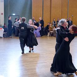 Tanzenden Paare in eleganten Kleidern auf einer Holz-Tanzfl&auml;che w&auml;hrend einer Tanzveranstaltung im Hintergrund.