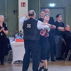Tanzpaar in eleganter Kleidung beim Tanzen, umgeben von applaudierendem Publikum in einer festlichen Halle.