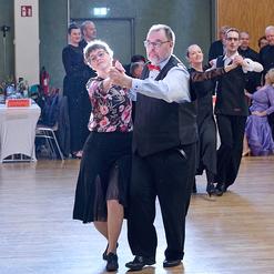 Tanzpaar in eleganter Kleidung vollf&uuml;hrt einen Langsame-Walzer in einem festlichen Saal mit Publikum im Hintergrund.