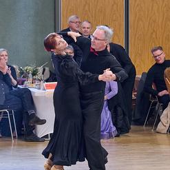 Tanzpaar in eleganter Kleidung f&uuml;hrt einen Tango auf einer Tanzfl&auml;che, Zuschauer applaudieren im Hintergrund.