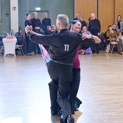 Tanzendes Paar in eleganter Kleidung, umgeben von Zuschauern in einem festlichen Saal.