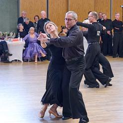 Tanzpaar f&uuml;hrt einen engagierten Tango auf einer Tanzfl&auml;che, Zuschauer beobachten im Hintergrund.