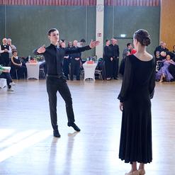 Tanzpaar auf einer Fl&auml;che, Mann in schwarzem Outfit dirigiert Frau in schwarzem Kleid vor Publikum in festlicher Kleidung.