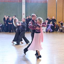 Tanzpaar in eleganter Kleidung tanzt in einem hellen Saal, umgeben von Zuschauern an festlich gedeckten Tischen.