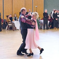 Tanzendes Paar auf einer Veranstaltung, umgeben von Zuschauern in einem gro&szlig;en Saal mit Holzfu&szlig;boden.