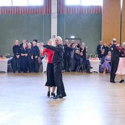 Tanzende Paare in eleganter Kleidung auf einer Tanzfl&auml;che, Zuschauer sitzen an Tischen im Hintergrund.