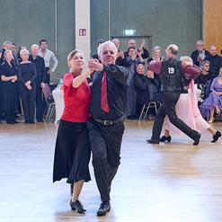Tanzendes Paar in eleganter Kleidung auf einer Tanzfl&auml;che, Publikum beobachtet und applaudiert im Hintergrund.