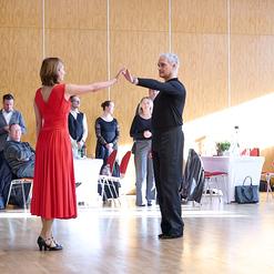 Tanzpaar in eleganter Kleidung f&uuml;hrt einen Schritt auf einer Tanzfl&auml;che, Zuschauer beobachten im Hintergrund.