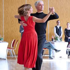 Tanzpaar in eleganter Kleidung f&uuml;hrt einen Walzer in einem festlichen Saal auf. Tische im Hintergrund sind dekoriert.