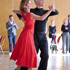 Tanzendes Paar in eleganter Kleidung, sie in rotem Kleid, er in schwarzem Oberteil, auf einem Parkettboden.