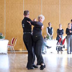 Tanzpaar f&uuml;hrt einen Schritt auf einer Tanzfl&auml;che, Zuschauer im Hintergrund beobachten die Vorf&uuml;hrung.