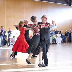 Tanzpaar in eleganter Kleidung f&uuml;hrt einen Tanz auf einer Holzbodenfl&auml;che, Zuschauer applaudieren im Hintergrund.