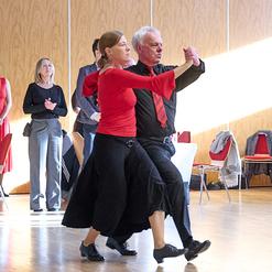 Tanzendes Paar in formeller Kleidung, umgeben von Zuschauern in einem hellen Saal mit Holzverkleidung.
