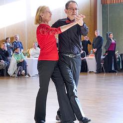 Tanzpaar in eleganter Kleidung tanzt miteinander auf einer hell erleuchteten Tanzfl&auml;che, Zuschauer im Hintergrund.