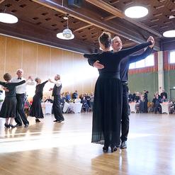 Tanzveranstaltung in einem Saal mit Paaren, die in eleganter Kleidung auf Holzfu&szlig;boden tanzen. Publikum schaut zu.