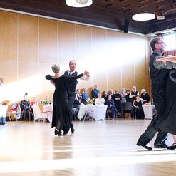 Paare tanzen in eleganter Kleidung auf einer Holzparkettfl&auml;che, w&auml;hrend Zuschauer in einem Festsaal zuschauen.