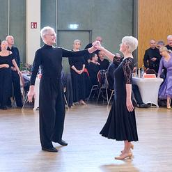 T&auml;nzerpaar in eleganter Kleidung tanzt auf einem Parkettboden, umgeben von Zuschauern bei einer Tanzveranstaltung.