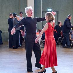 Tanzpaar in Aktion: Mann in Schwarz und Frau in rotem Kleid tanzen auf einem Parkettboden, im Hintergrund weitere T&auml;nzer.