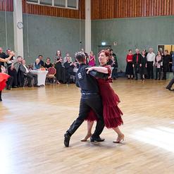 Tanzpaar in stilvoller Kleidung tanzt auf einer Holzfl&auml;che, umgeben von Zuschauern und weiteren T&auml;nzern.
