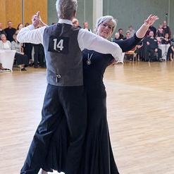 Tanzpaar in eleganter Kleidung f&uuml;hrt einen Walzer auf einer Tanzfl&auml;che, Publikum im Hintergrund beobachtet.