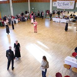 Tanzveranstaltung in einer Halle mit T&auml;nzern in eleganter Kleidung und Publikum an Tischen im Hintergrund.