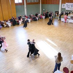 Tanzsportturnier im Saal mit t&auml;nzerischen Paaren und Publikum an Tischen, im Hintergrund eine Band und eine Banner.