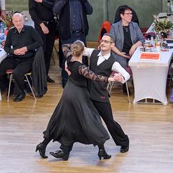 Tanzpaar in eleganter Kleidung tanzt leidenschaftlich auf einer Tanzfl&auml;che, umgeben von Zuschauern an festlich gedeckten Tischen.