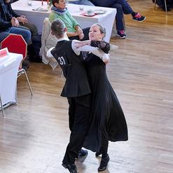 Tanzendes Paar in eleganter Kleidung auf einer Festveranstaltung, Zuschauer beobachten aufmerksam im Hintergrund.