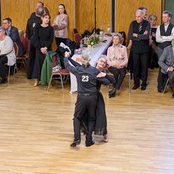 Tanzendes Paar in eleganter Kleidung, umgeben von Zuschauern in einem festlichen Saal.