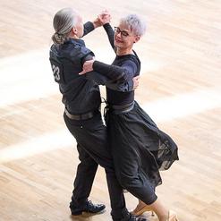 Tanzpaar in eleganter Kleidung, das sich auf einem hellen Parkettboden im Cha-Cha bewegt.