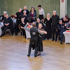 Tanzpaar in eleganter Kleidung w&auml;hrend eines Wettbewerbs, umgeben von zuschauenden G&auml;sten an festlich gedeckten Tischen.