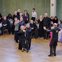 Tanzpaar in eleganter Kleidung auf einer Tanzveranstaltung, umgeben von Zuschauern an festlich gedeckten Tischen.