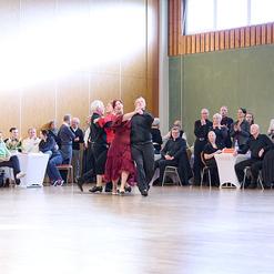 Tanzendes Paar in festlicher Kleidung auf einer Tanzfl&auml;che, umgeben von Zuschauern, die aufmerksam beobachten.
