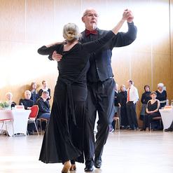 Tanzendes Paar in eleganter Kleidung auf einer Tanzfl&auml;che bei einer Veranstaltung, umgeben von sitzendem Publikum.