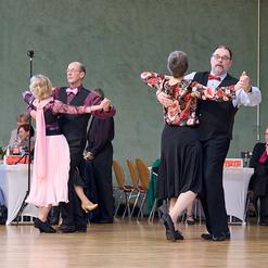 Tanzpaare in eleganter Kleidung bei einem Tanzwettbewerb auf einer Holzfl&auml;che, Publikum im Hintergrund.