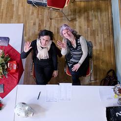 Zwei Frauen sitzen an einem Tisch, winken freundlich und umgeben von Dekoration und Unterlagen.