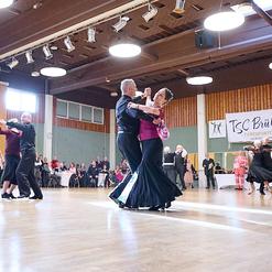 Tanzpaare in eleganter Kleidung tanzen in einem gro&szlig;en Saal mit Tischanordnung im Hintergrund. Banner des TSC Br&uuml;hl sichtbar.