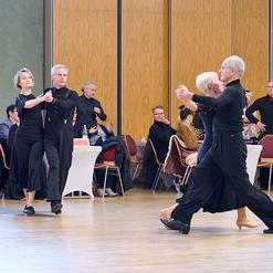 Tanzende Paare in schwarzer Kleidung auf einer Tanzfl&auml;che, umgeben von Zuschauern in einem festlichen Saal.