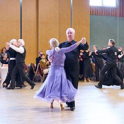 Tanzpaar in eleganter Kleidung tanzt in einem festlichen Saal, Zuschauer applaudieren im Hintergrund.