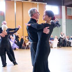 Tanzpaar in eleganter Kleidung bei einer Tanzveranstaltung auf einer Holzb&uuml;hne, umgeben von G&auml;sten.