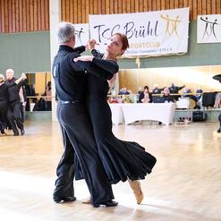 Tanzpaar in eleganter Kleidung tanzt in einem Turnier, Hintergrund mit Zuschauern und Tisch.