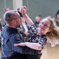 Tanzpaar in eleganter Kleidung tanzt l&auml;chelnd auf einer Tanzfl&auml;che, im Hintergrund Publikum applaudiert.