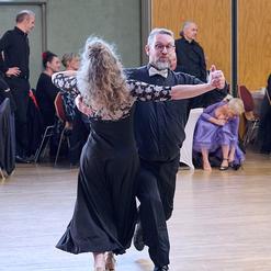 Tanzendes Paar in eleganter Kleidung, umgeben von G&auml;sten in einem festlichen Saal.