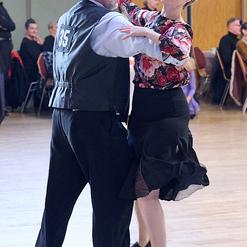 Tanzpaar in eleganter Kleidung beim Tanzen auf einer Tanzfl&auml;che, umgeben von weiteren G&auml;sten im Hintergrund.