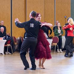 Tanzpaar in eleganter Kleidung, im Mittelpunkt eines Tanzsaals, umgeben von Zuschauern an Tischen.