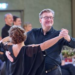 Tanzpaar in formeller Kleidung tanzt l&auml;chelnd in einem festlich dekorierten Saal. Menschen im Hintergrund beobachten das Geschehen.